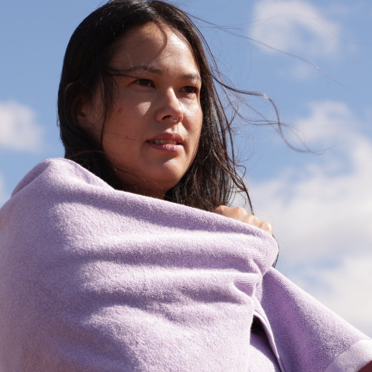 woman with brunette hair wrapped in a lilac bath towel with blue sky behind her